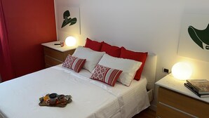 A neatly made bed with red and white pillows, a bedside table with a lamp, and a small clock. - [5 min from Uffizi] Lady Suite in Center, Wifi, Comfort (FLORENCE)