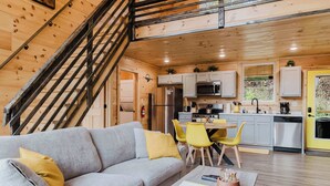 Interior de una cabaña moderna con una escalera de madera, un sofá gris con cojines amarillos, un área de comedor con sillas amarillas y una cocina con armarios blancos y electrodomésticos de acero inoxidable.