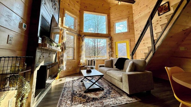 Interior acogedor de una cabaña con chimenea, un sofá, una mesa de centro y grandes ventanales que ofrecen vistas al exterior.
