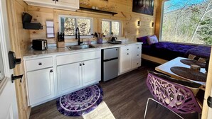 Une cuisine compacte équipée d’un évier, d’une cuisinière et de placards, un coin repas avec une chaise violette, et un lit donnant sur la forêt.