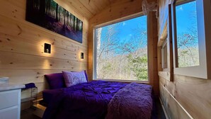 Une chambre dans une cabane en bois, avec un lit, une literie violette, une fenêtre donnant sur les arbres et une lampe fixée au mur.