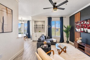 Un salon moderne avec un canapé, une table basse et un téléviseur. On y trouve également un ventilateur de plafond, un coin repas avec une table et des chaises, ainsi qu'une cuisine équipée de meubles de rangement.