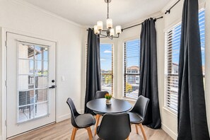 A dining area with a round table, four chairs, a chandelier, and large windows with blinds.