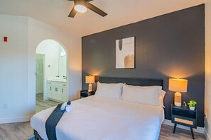 A hotel room with a bed, two bedside tables with lamps, a ceiling fan, a large painting on the wall, and an arched doorway leading to a bathroom.