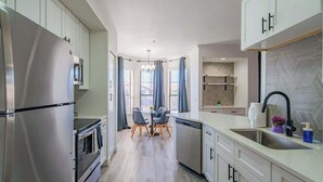 A modern kitchen with stainless steel appliances, a dining area with a table and chairs, and a view of the outside through large windows.