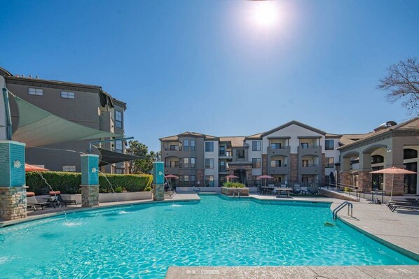 A swimming pool with a clear blue water surface, surrounded by multi-story buildings with balconies, and a clear sky above.