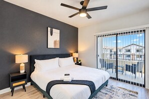 A bedroom with a bed, bedside lamps, a ceiling fan, a window with a view, and a painting on the wall.