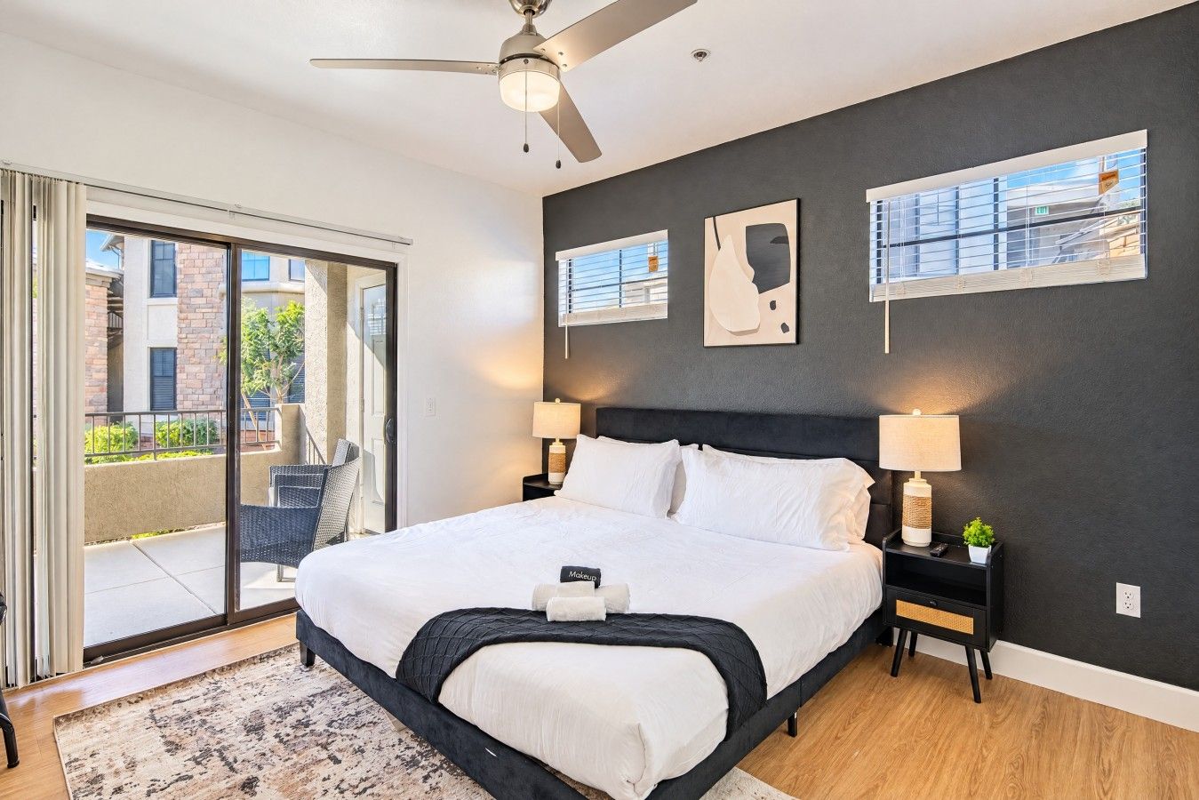 A modern bedroom with a large bed, a ceiling fan, and a view through the glass door to the outside.