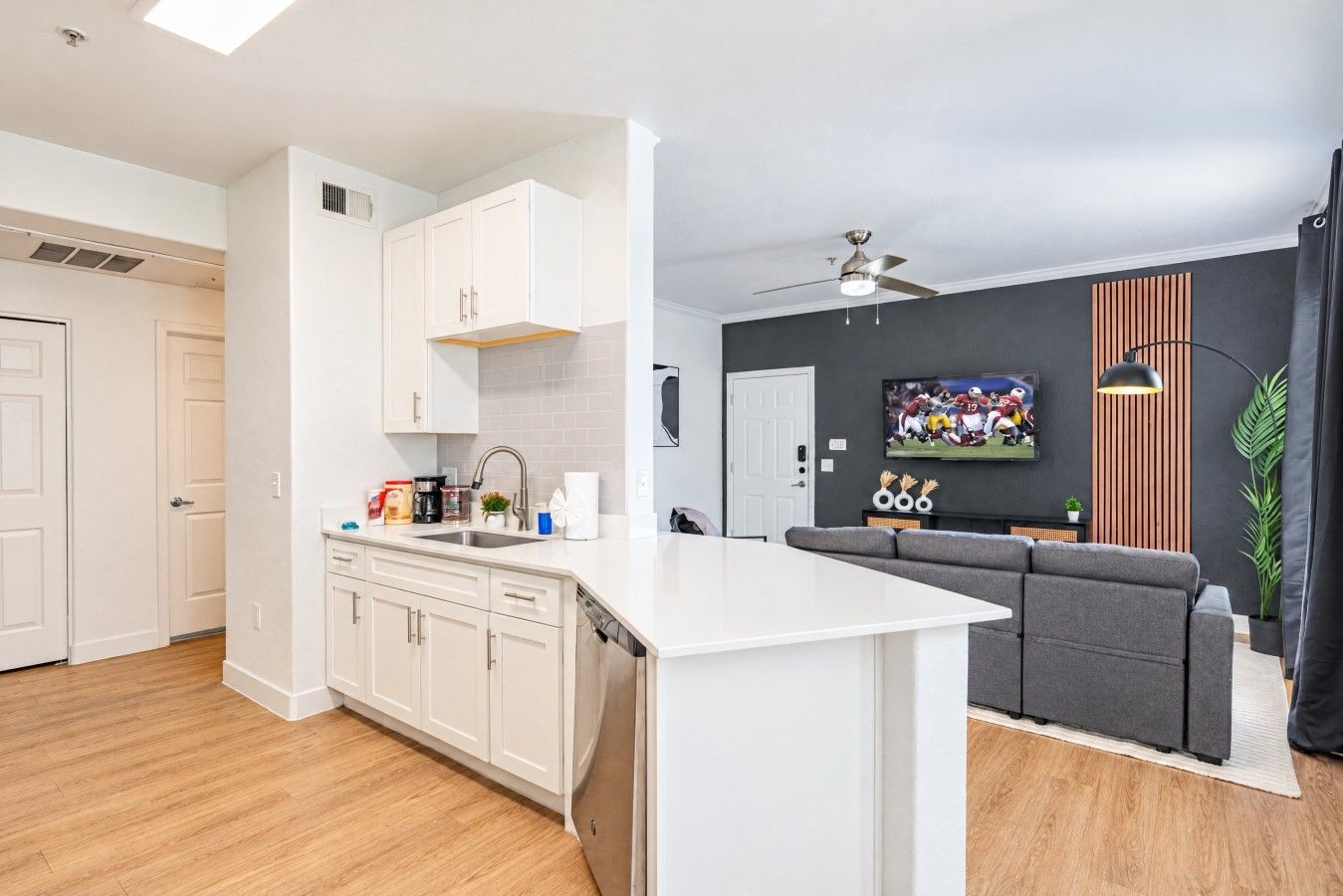 A modern kitchen with white cabinets, a built-in dishwasher, and a grey sofa in the living area.