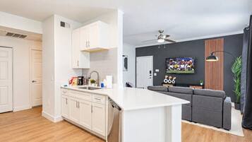 A modern kitchen with white cabinets, a built-in dishwasher, and a grey sofa in the living area.
