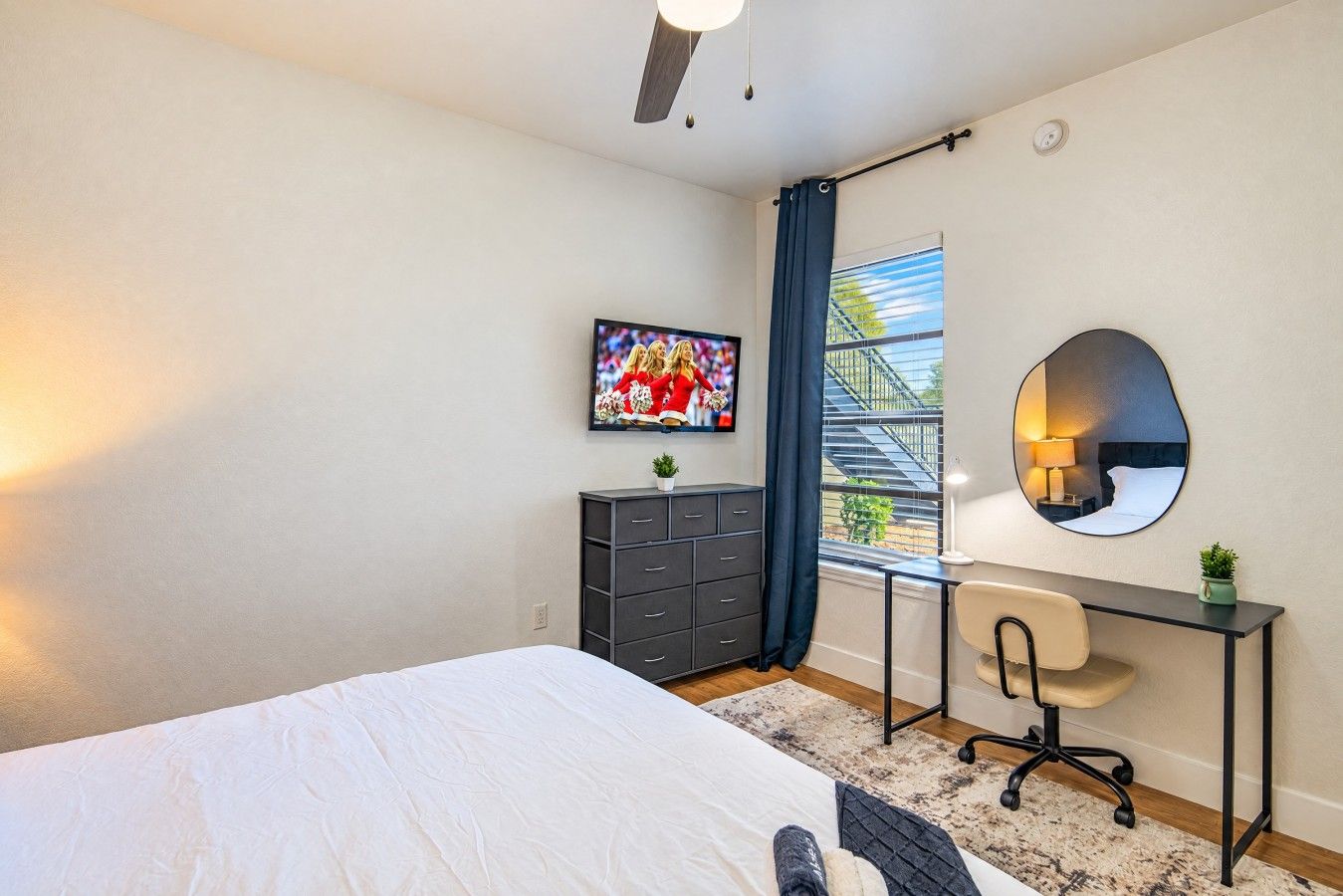Un dormitorio con cama, escritorio, silla, cómoda, ventana con vistas y espejo.