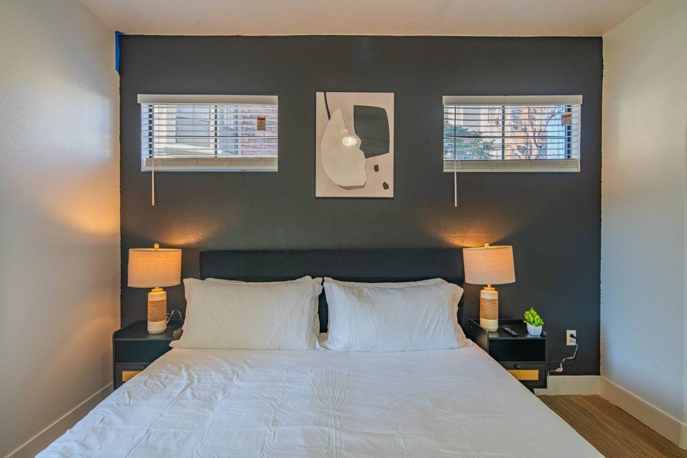 Een slaapkamer met een bed, twee bedlampen, een nachtkastje met een plant en twee ramen met zonwering.