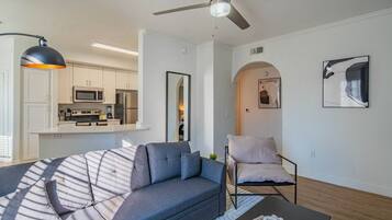 Un salon moderne avec un canapé, une table basse et un fauteuil. On aperçoit à l’arrière-plan un coin cuisine avec des placards et des appareils électroménagers.