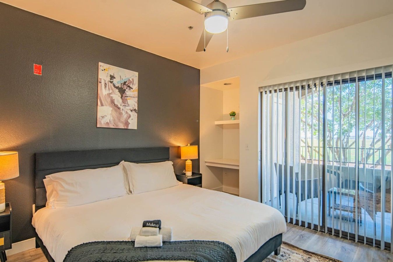 A hotel room with a bed, two bedside lamps, a ceiling fan, a painting on the wall, and a view through the window.