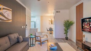A modern living room with a sofa, armchair, coffee table, and a TV. There is a plant and artwork on the walls. - Near the Stadium with  Pool Gym Parking HotTub WD (Glendale)