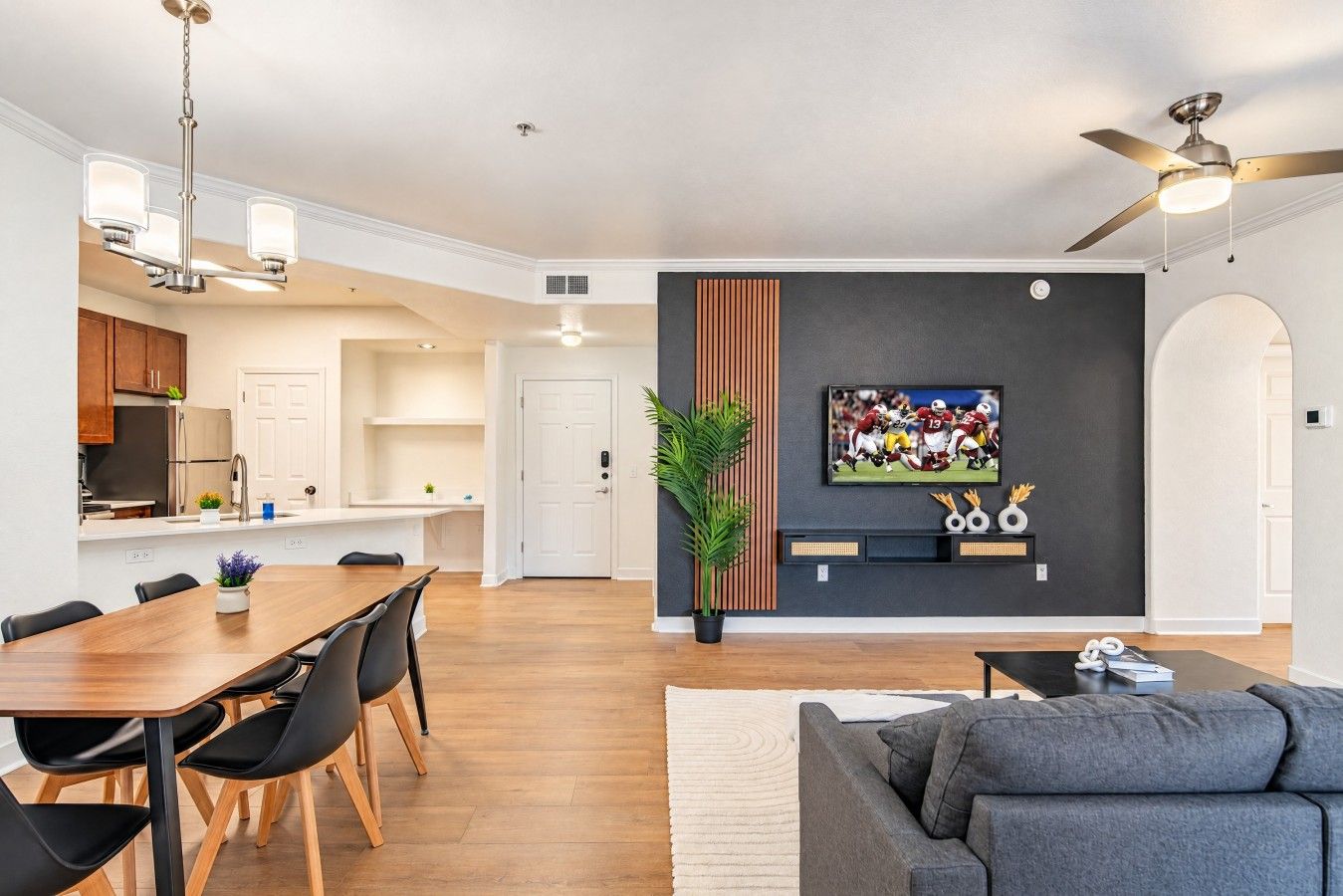 A modern kitchen with white cabinets, a dining area with a wooden table and black chairs, a gray sofa, and a TV mounted on a dark wall.