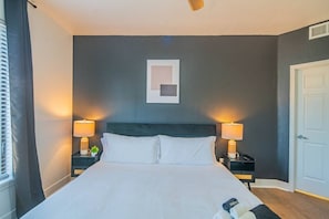 A hotel room with a bed, two bedside tables with lamps, a door, and a window with blinds.