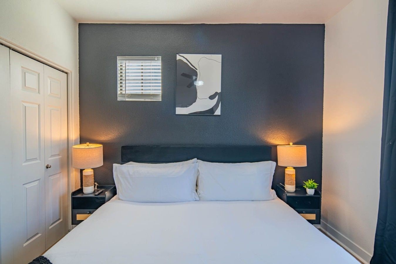 A hotel room with a bed, two bedside lamps, a window with blinds, and a wall with two framed pictures.