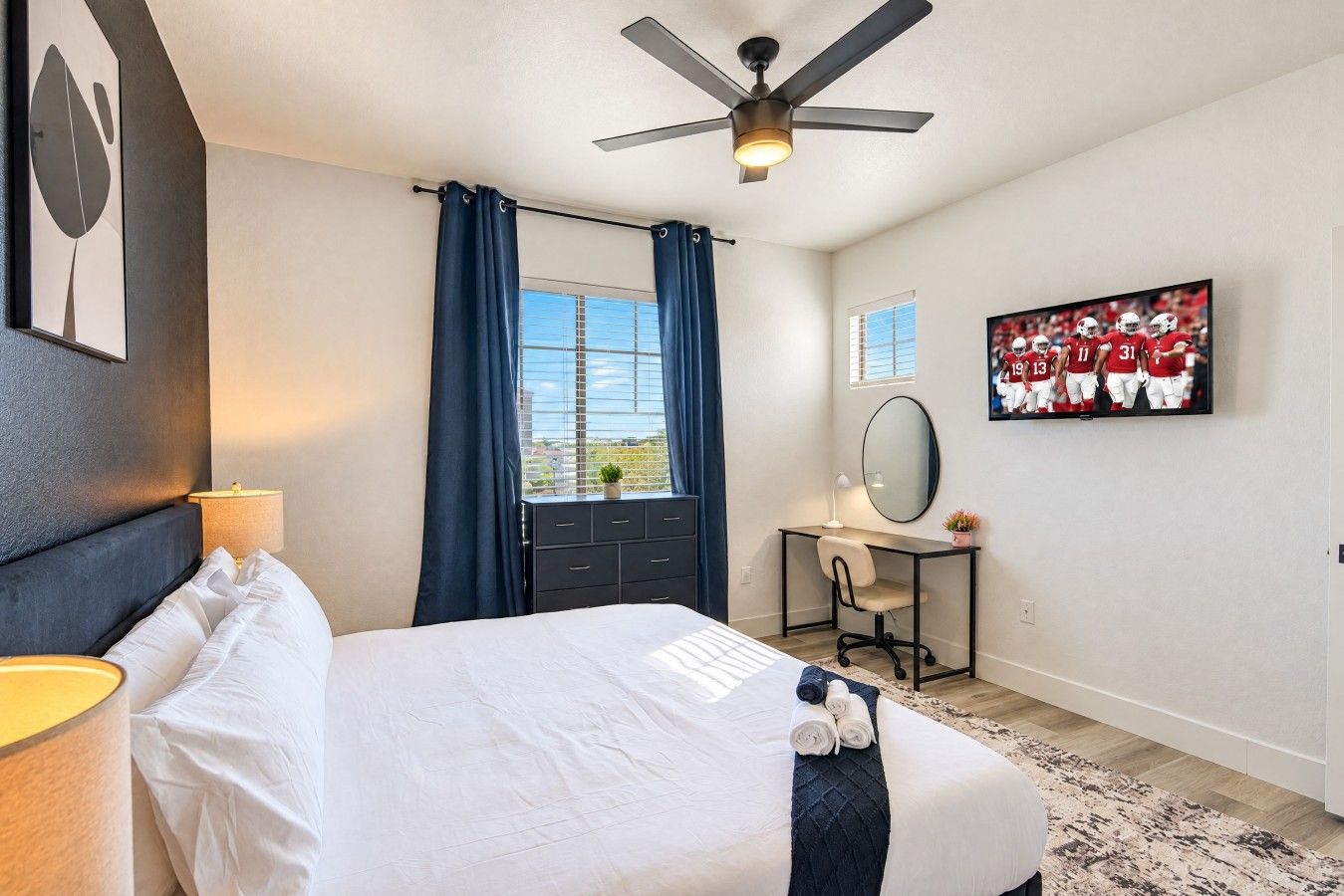 Une chambre d’hôtel comprenant un lit, un bureau, une chaise, un ventilateur de plafond, une fenêtre avec des rideaux, un miroir fixé au mur et un tableau encadré accroché au mur.