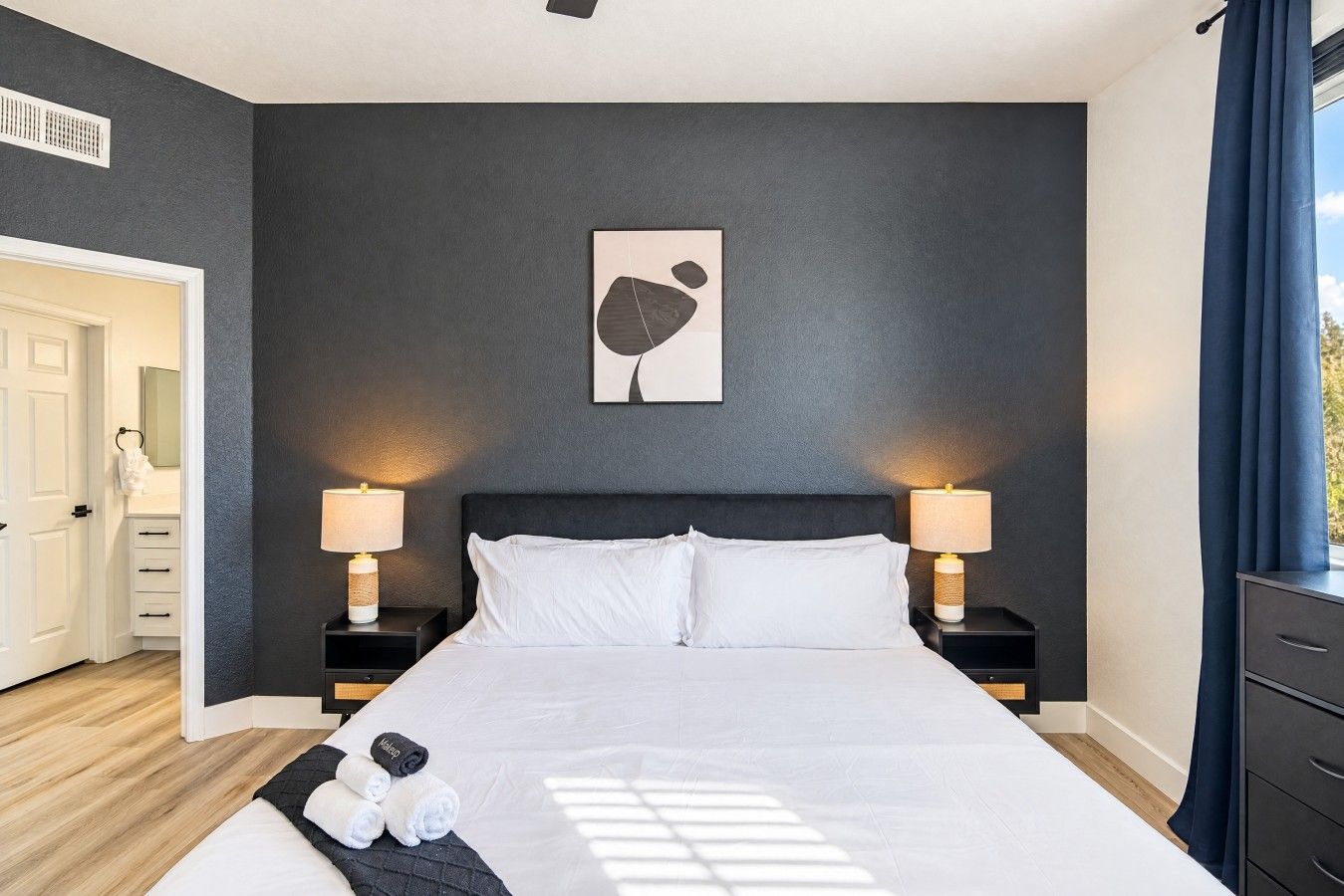 A modern bedroom with a large bed, two bedside tables with lamps, a dark wall, and a single artwork above the bed.
