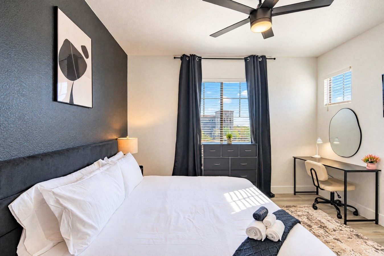 Une chambre d’hôtel moderne équipée d’un lit, d’un bureau, d’une chaise, d’un ventilateur de plafond, d’une fenêtre avec des rideaux et d’un miroir fixé au mur.