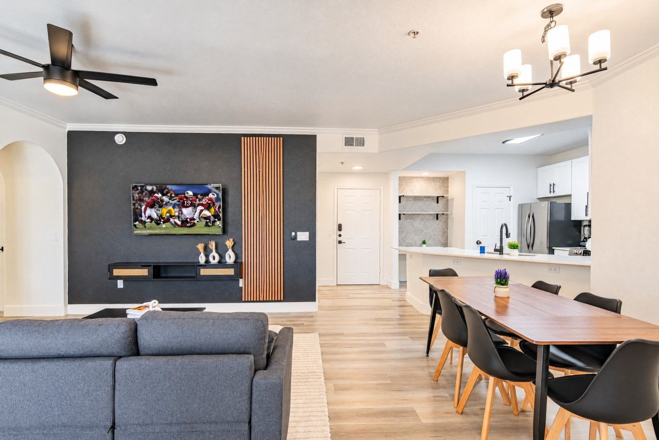 A modern living room with a grey sofa, a dining area with a wooden table and black chairs, a kitchen with white cabinets, and a ceiling fan.