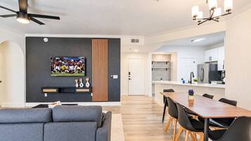 A modern living room with a grey sofa, a dining area with a wooden table and black chairs, a kitchen with white cabinets, and a ceiling fan.