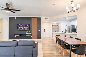 Uma sala de estar moderna com um sofá cinzento, um espaço de refeições com mesa de madeira e cadeiras pretas, uma cozinha com armários brancos e um ventilador de teto.