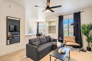 A modern living room with a sectional sofa, a flat-screen TV, a ceiling fan, and a dining area in the background.