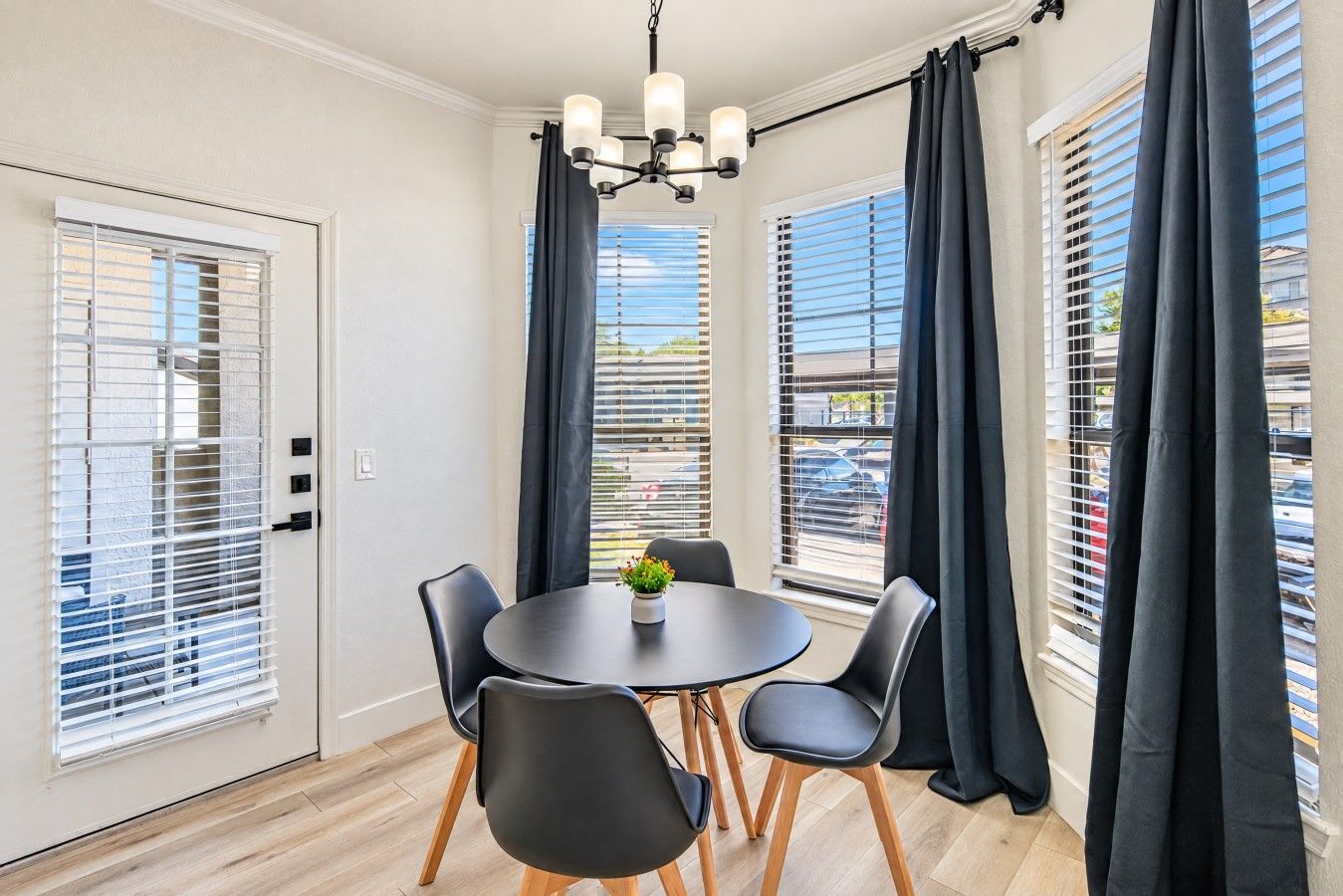 A dining area with a round table, four chairs, a chandelier, and large windows with blinds.