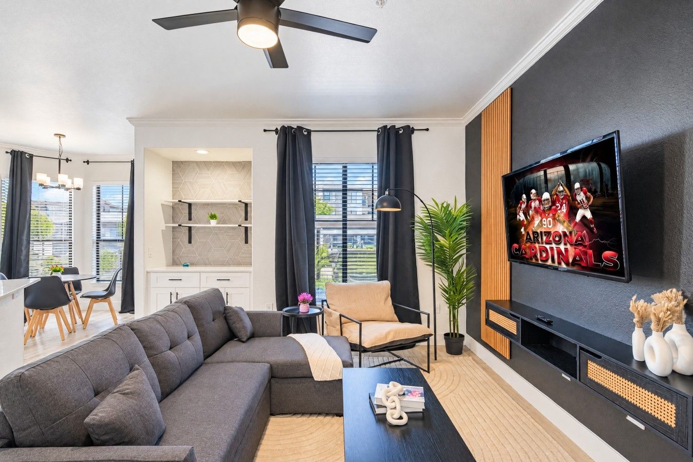A modern living room with a sectional sofa, a dining area, a kitchen, and a TV displaying the Arizona Cardinals.