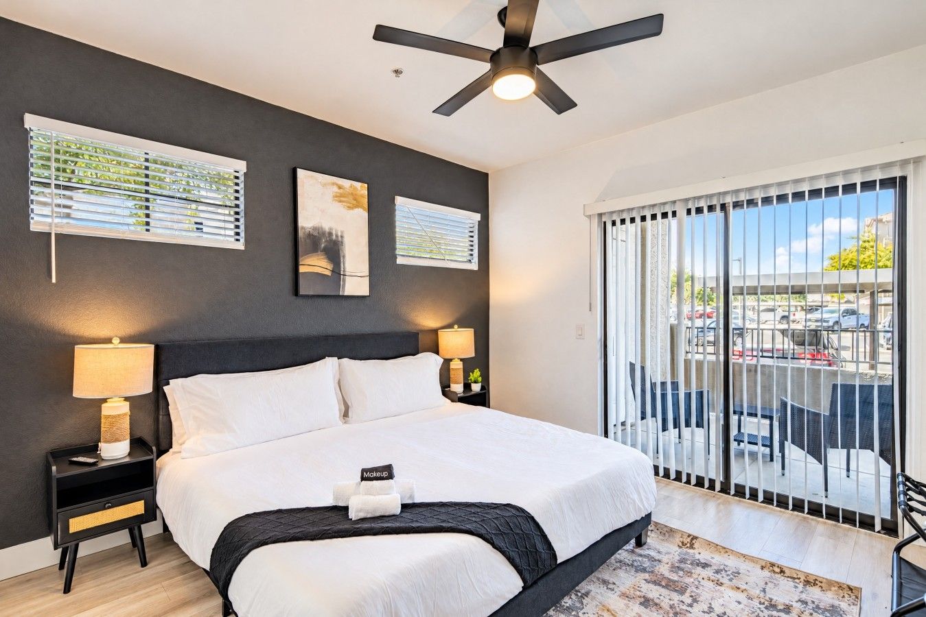 A hotel room with a bed, two bedside lamps, a ceiling fan, and a view of a parking area through large windows.