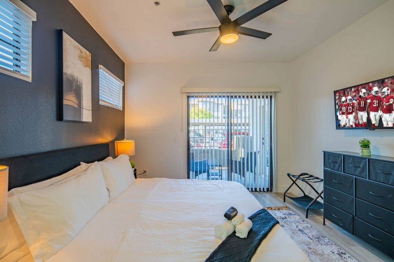A hotel room with a bed, a ceiling fan, a TV, a dresser, and a window with blinds.