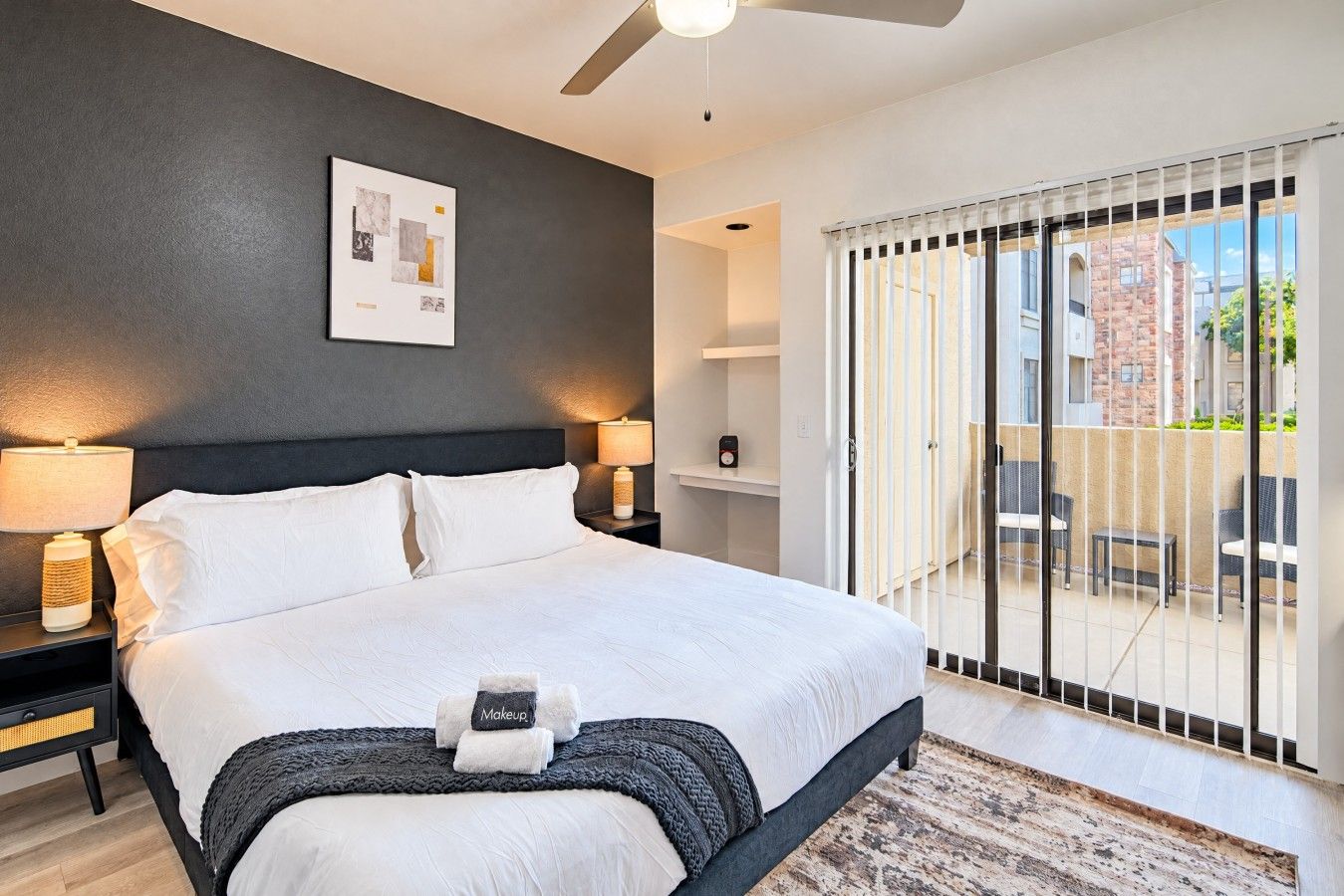Une chambre d’hôtel moderne avec un grand lit, deux tables de chevet avec des lampes, un ventilateur de plafond, une vue sur le balcon extérieur et un tableau encadré au mur.