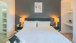 Quarto de hotel com uma cama grande, duas luminárias de cabeceira, uma obra de arte na parede e uma mesinha de cabeceira com uma planta.