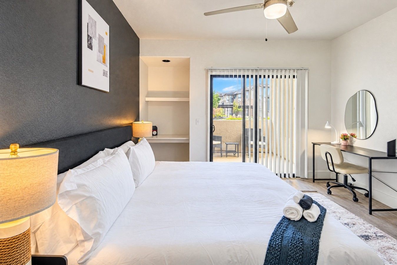 A hotel room with a bed, a desk, a chair, a lamp, a ceiling fan, and a view of the outside.