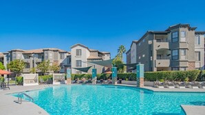 Piscina com espreguiçadeiras e guarda-sóis, cercada por edifícios com varandas.