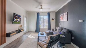 Uma sala de estar moderna com sofá, poltrona e mesa de centro. Há uma televisão montada na parede, um ventilador de teto e uma janela com cortinas.