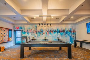 Una sala de billar con mesa de billar, dos pequeñas mesas y un mural abstracto y colorido en la pared.