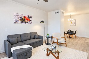 A modern living room with a grey sofa, a glass coffee table, and a dining area with a white chair and black chairs.