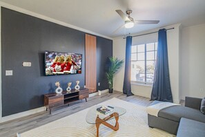 Una sala de estar moderna con una televisión de pantalla plana, un ventilador de techo, un sofá, una mesa de centro y un gran ventanal con cortinas.