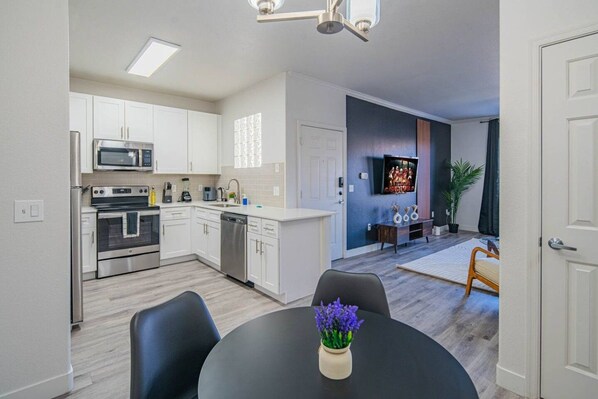 Una cocina moderna con armarios blancos, microondas, horno y lavavajillas. Hay una zona de comedor con mesa y sillas, y un televisor montado en la pared.