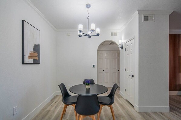 A dining area with a round table, four chairs, a chandelier, and a door leading to another room.