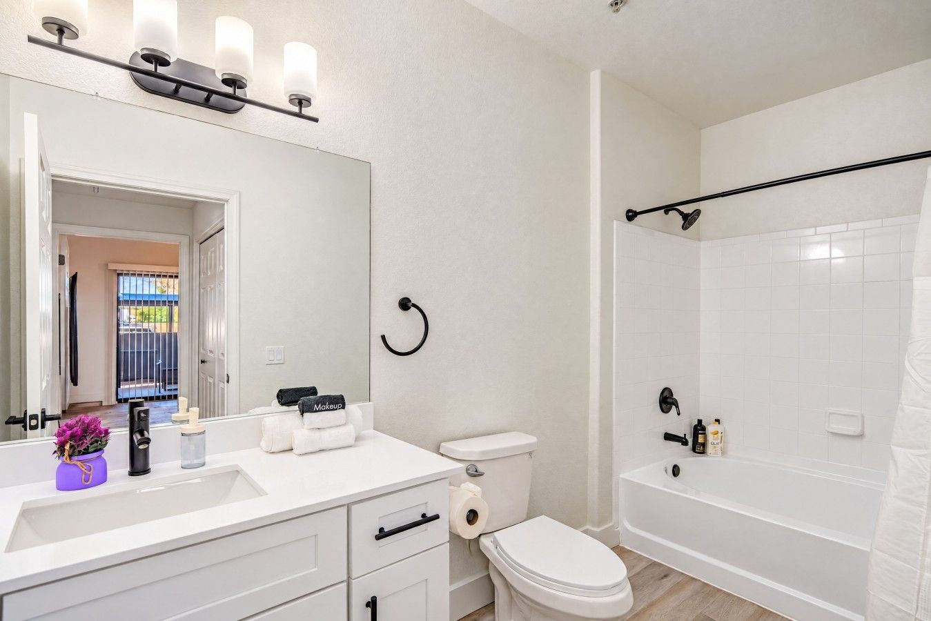 Un baño moderno con lavamanos, inodoro y bañera blancos. Hay un espejo con luces arriba y un perchero con toallas.