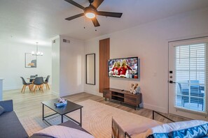 A modern living room with a flat-screen TV, a ceiling fan, a dining area with chairs, and a large window with blinds.