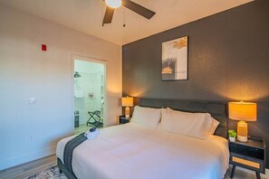 A hotel room with a bed, two bedside tables with lamps, a ceiling fan, a door leading to a bathroom, and a wall-mounted artwork.