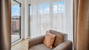 Un salon moderne avec un canapé et une grande fenêtre ornée de rideaux légers, offrant une vue sur les immebles et un balcon.