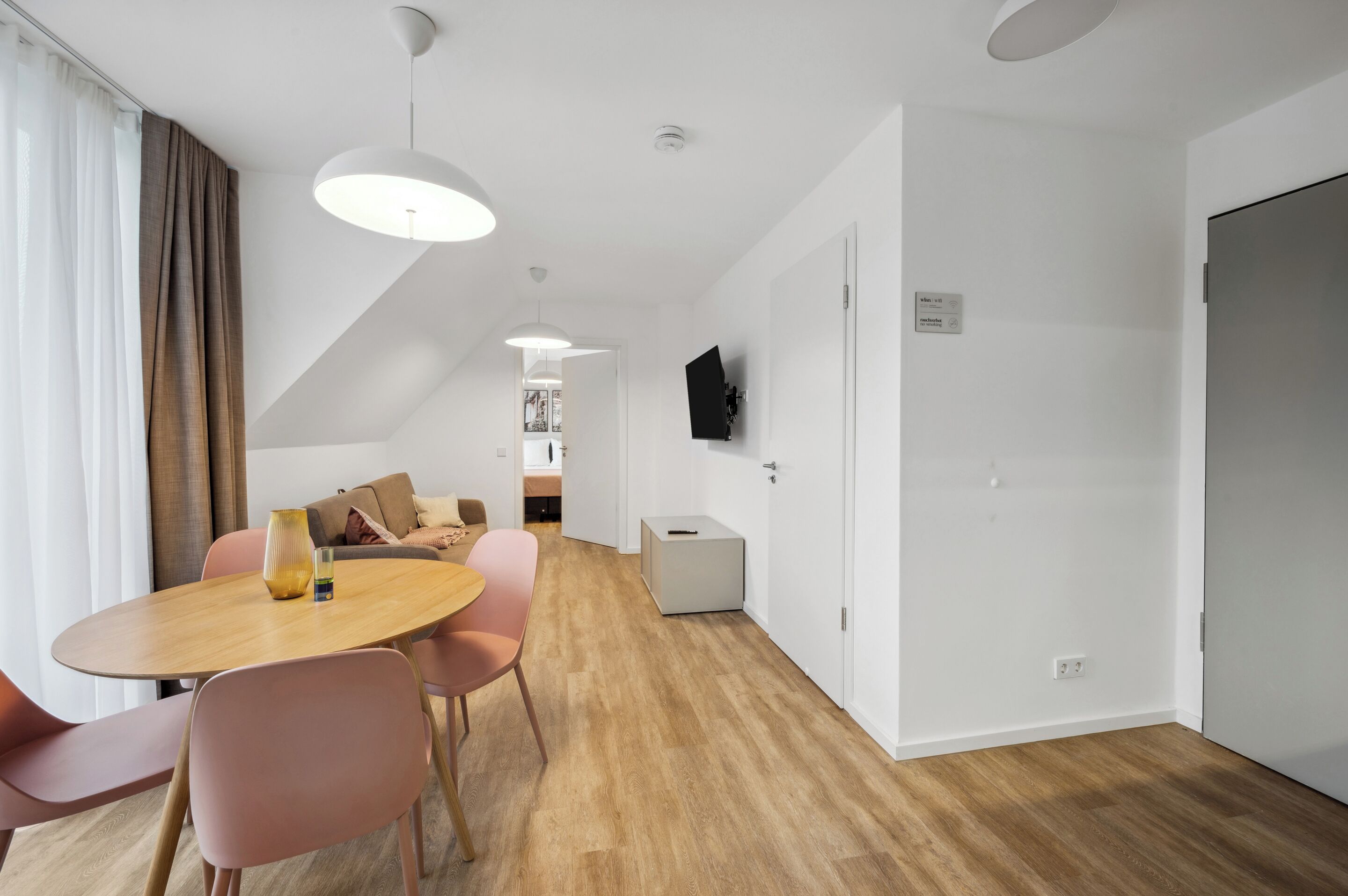 A modern hotel room with a wooden floor, a round dining table with pink chairs, a sofa with beige cushions, and a flat-screen TV mounted on the wall.