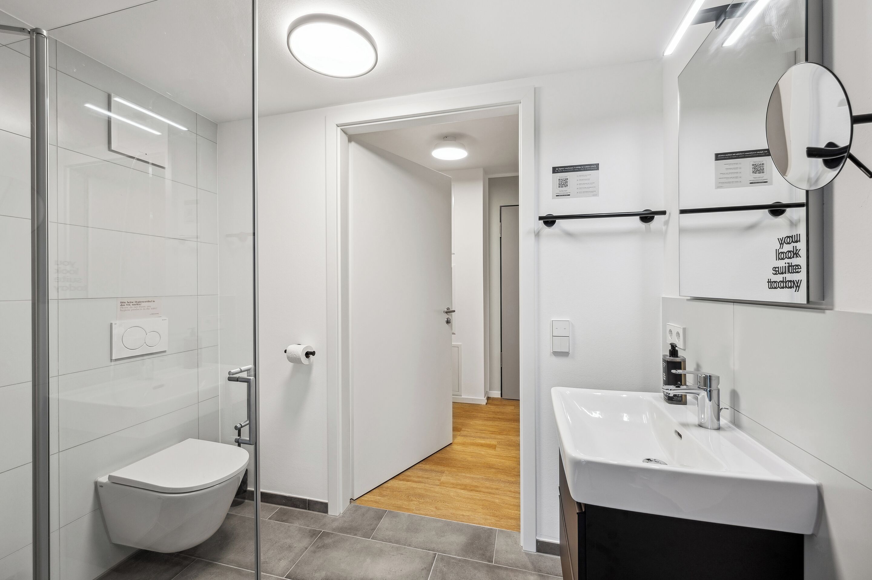 A modern bathroom with a glass shower enclosure, a white toilet, a sink with a mirror, and a wooden floor leading to a hallway.