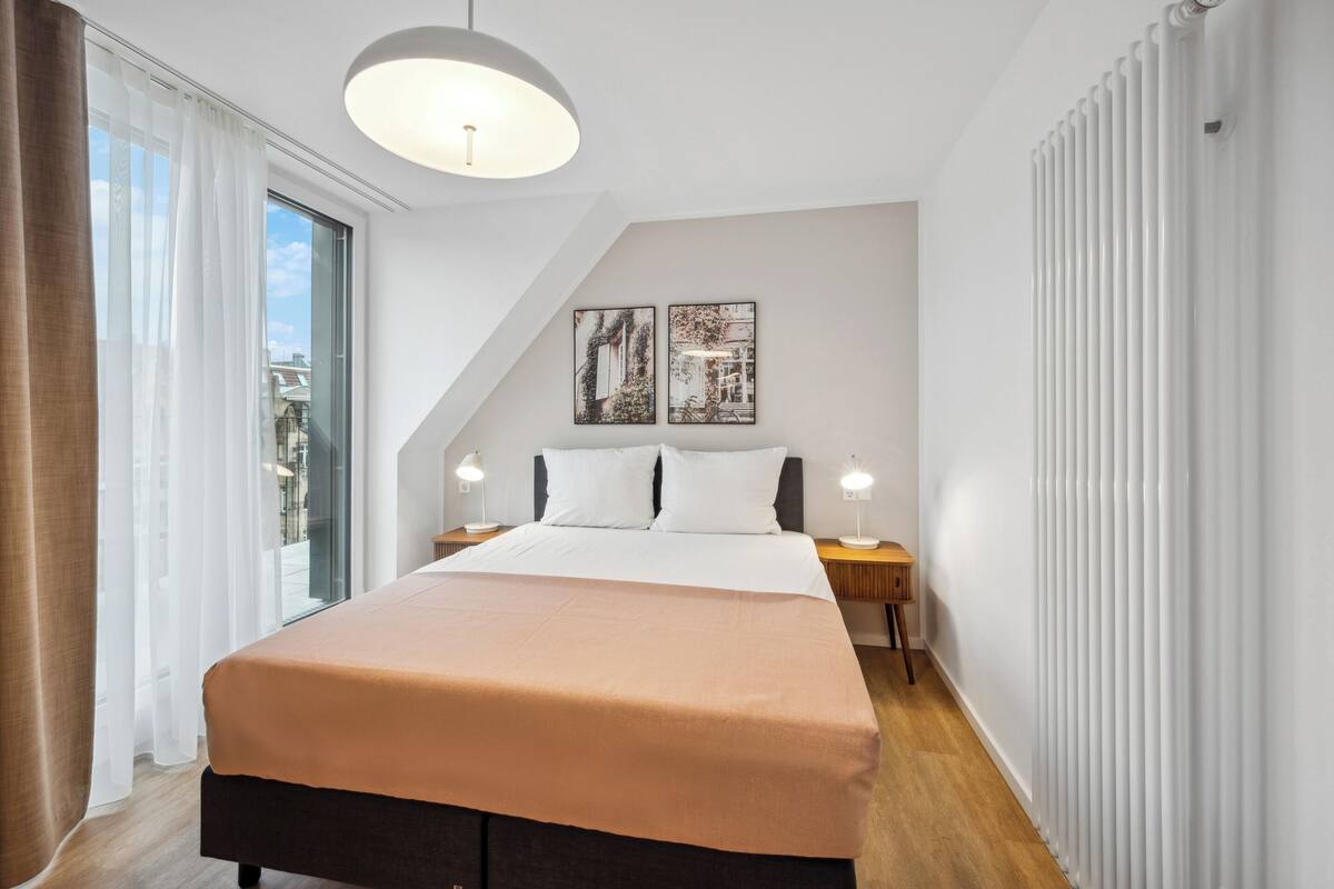 Une chambre d’hôtel moderne avec un grand lit, deux tables de chevet avec des lampes, un luminaire au plafond et une vue sur la ville par une grande fenêtre.